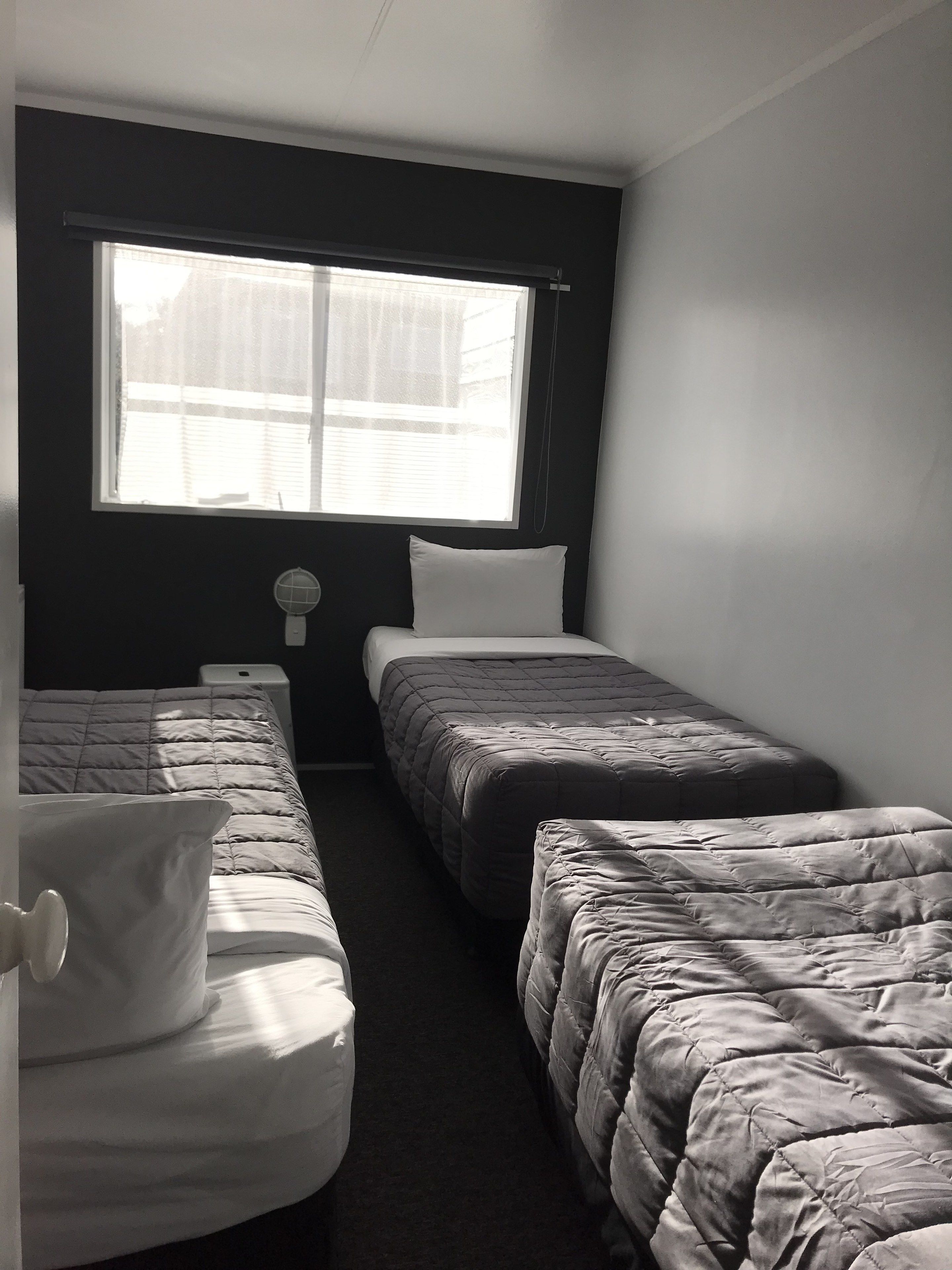 Ein Zimmer mit zwei Einzelbetten, einem Fenster mit Jalousien, einem kleinen Nachttisch mit Lampe und einer dunkel gestrichenen Wand.