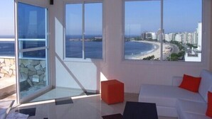 Un salón moderno con amplios ventanales que dan a una playa y al skyline de la ciudad.