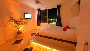 Habitación de hotel moderna con cama, ventilador de techo, televisión y vistas de la ciudad de noche.
