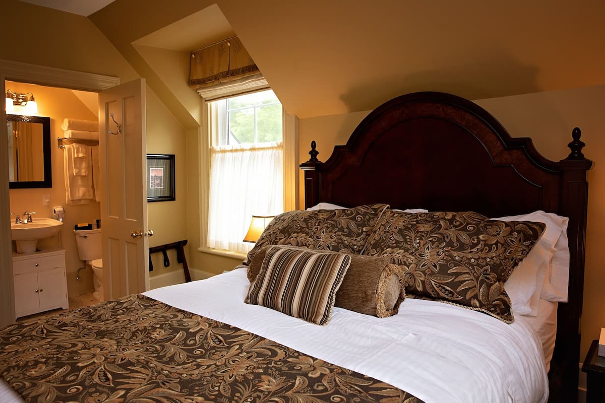 A bedroom with a large bed, a wooden headboard, a window with curtains, a bedside table, and a bathroom visible through an open door.