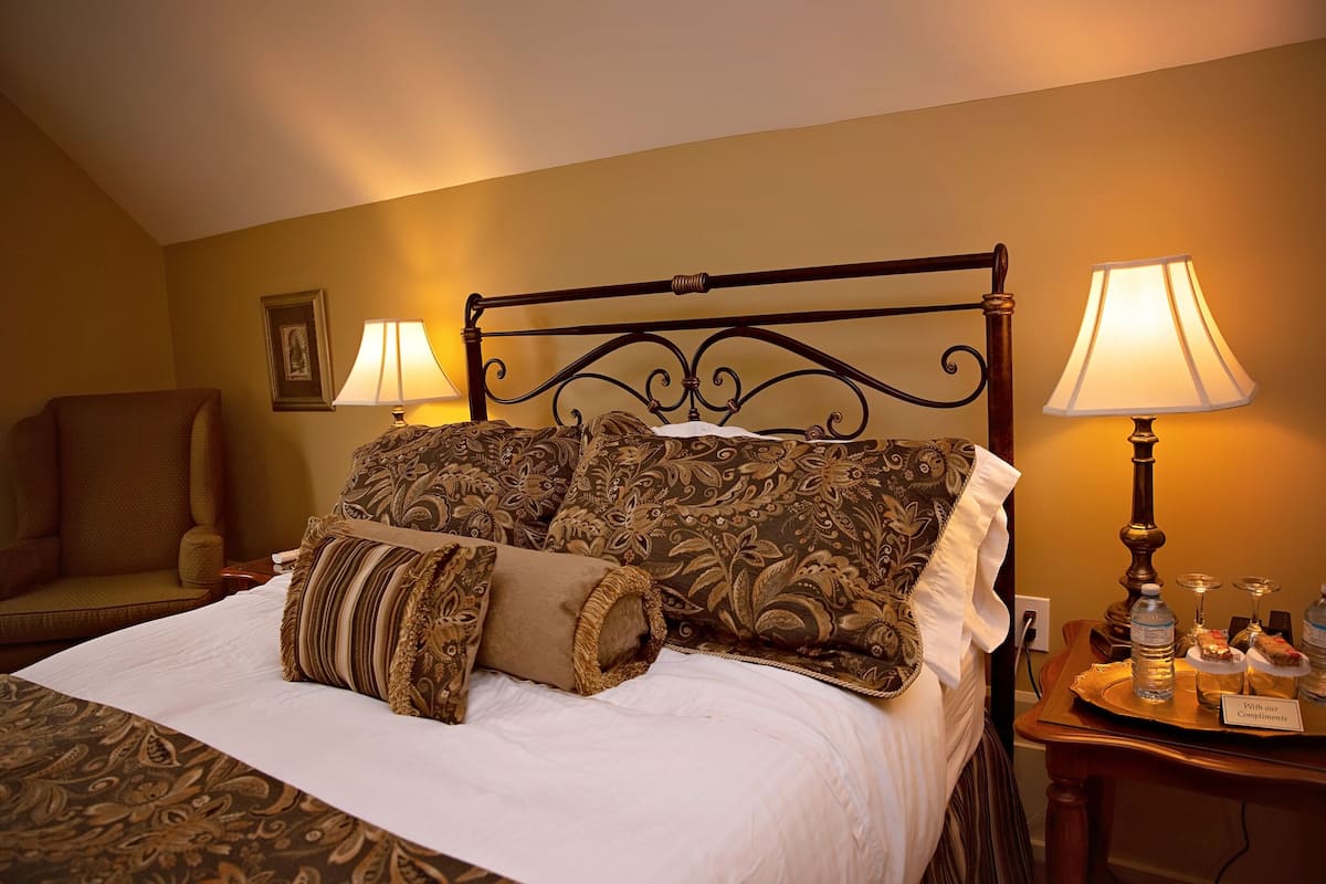 A neatly made bed with patterned bedding, a chair, and a nightstand with a lamp and a tray of snacks.