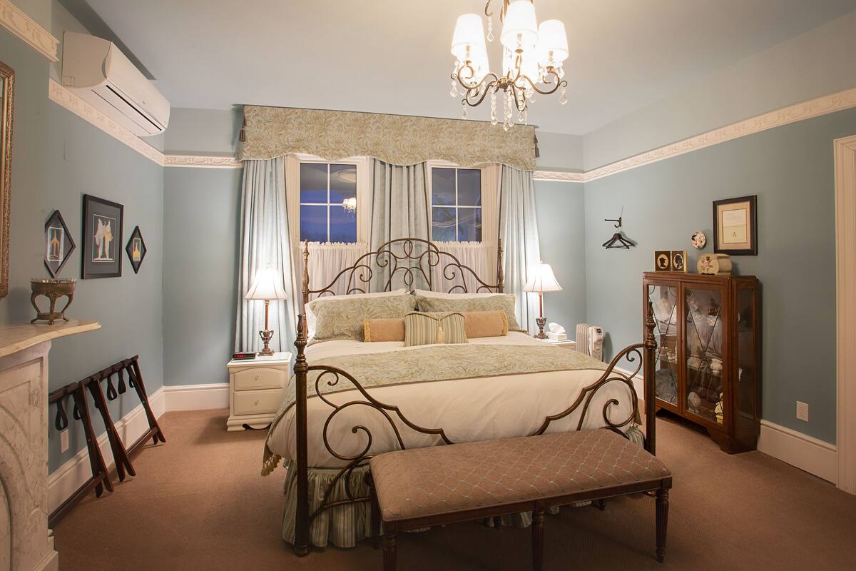 A bedroom with a four-poster bed, a chandelier, a bedside table, a dresser, and a bench.