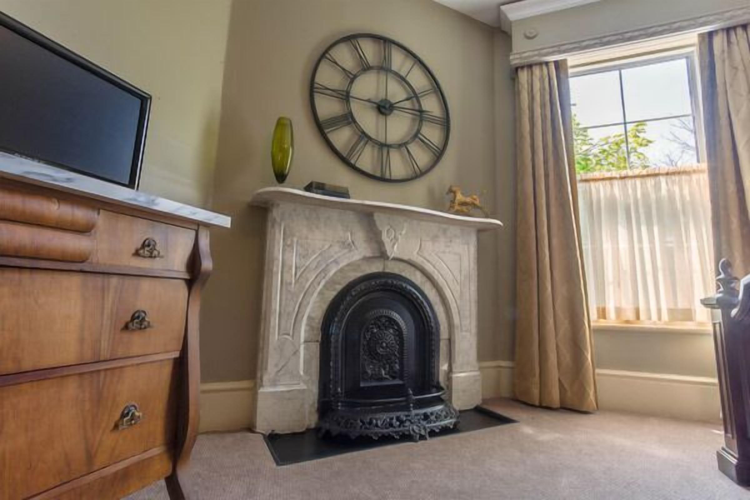 A room with a fireplace, a clock, a television on a cabinet, and a window with curtains.
