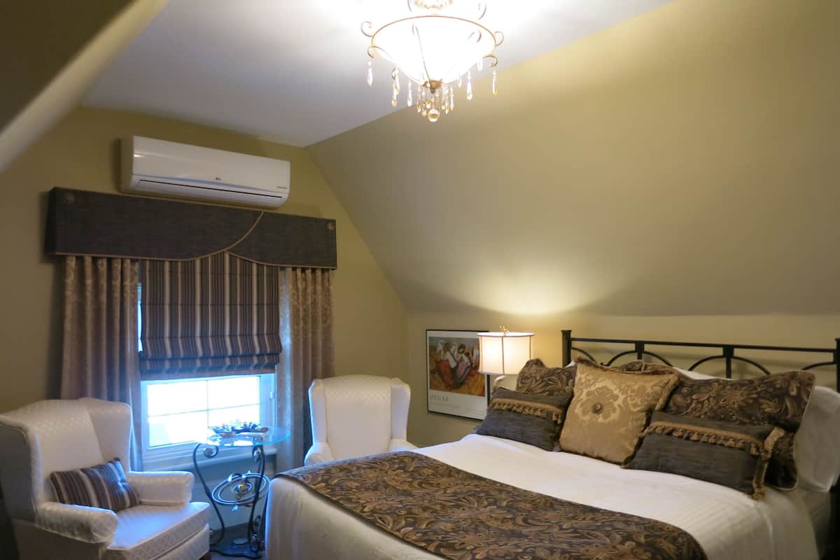 A bedroom with a bed, two armchairs, a table, a chandelier, an air conditioning unit, and a window with curtains.