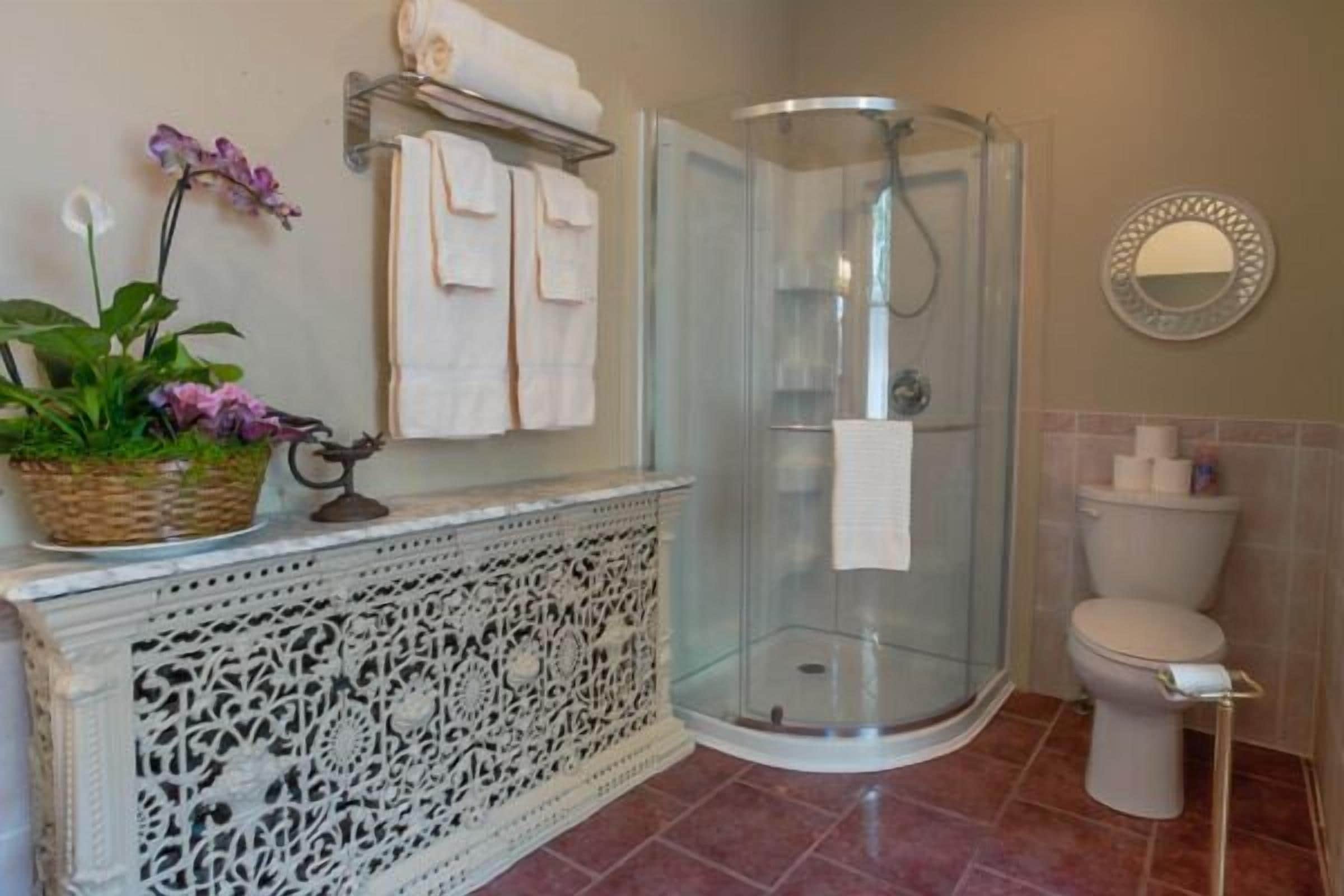 Un baño con cabina de ducha de vidrio, inodoro, un mueble decorativo con intrincados adornos y una cesta con flores.