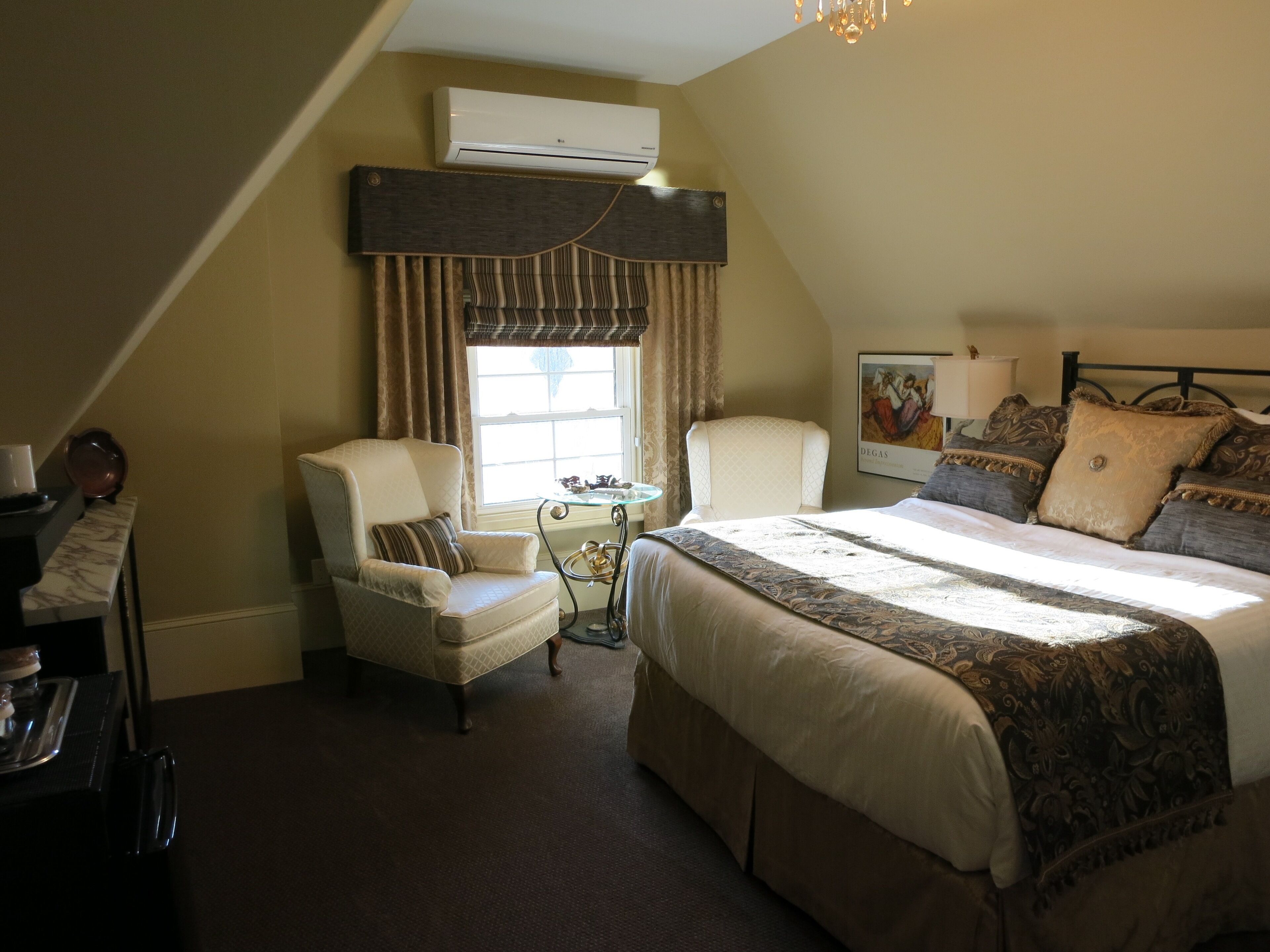 A bedroom with a large bed, two armchairs, a small table, and an air conditioning unit.