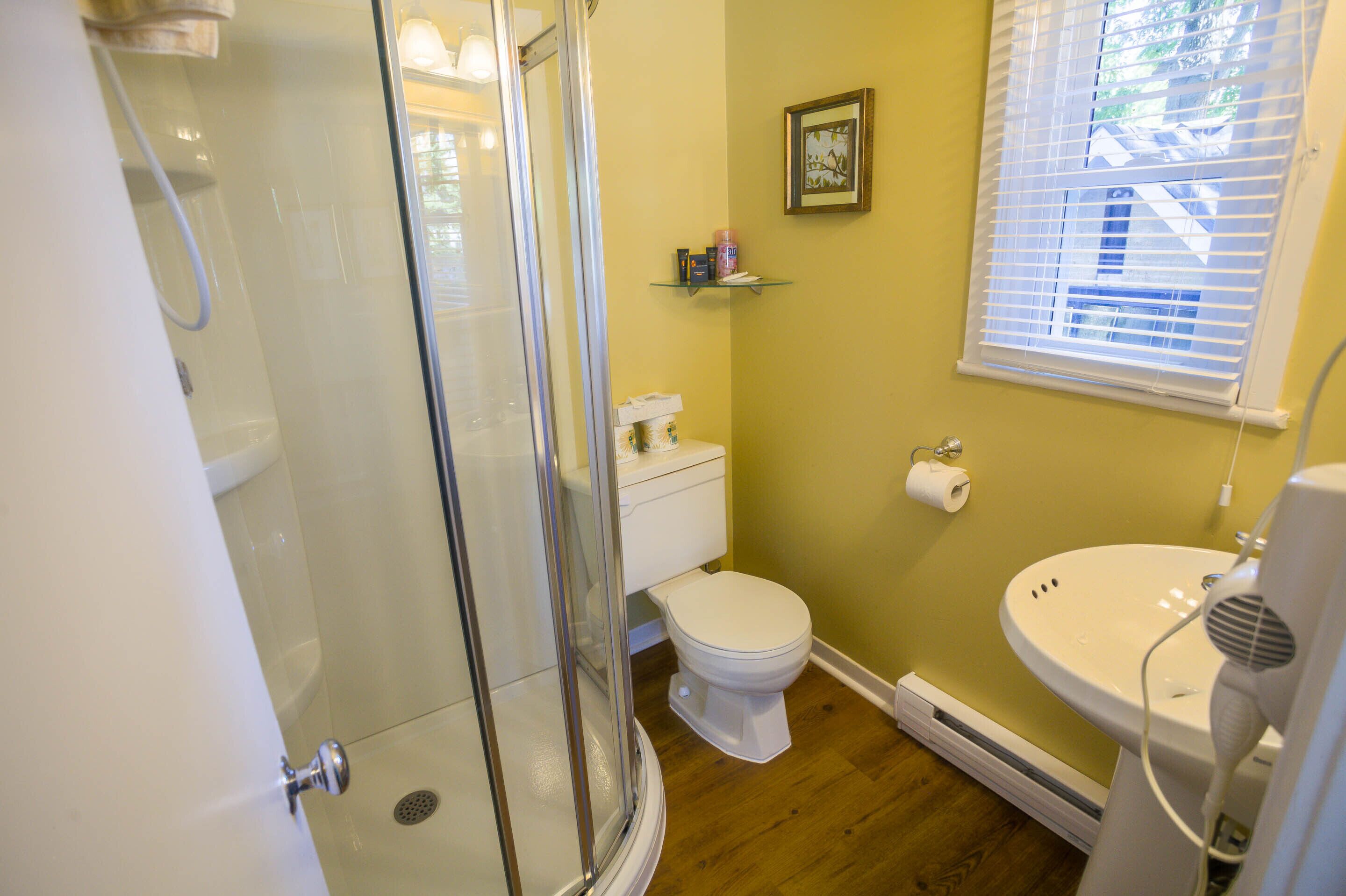 A bathroom with a glass-enclosed shower, a toilet, a sink, and a window with blinds.