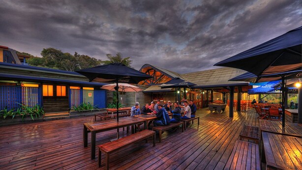 An outdoor dining area with wooden tables and benches, umbrellas, and a view of the building's exterior. - Fraser Island Retreat (K'gari (Fraser Island))
