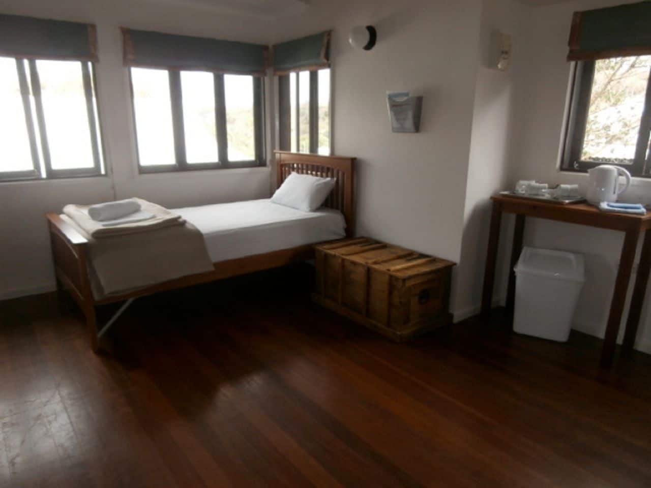 Ein kleines Zimmer mit einem Einzelbett, einem Holzkopfstück, einer hölzernen Kommode, einem kleinen Tisch mit Wasserkocher und einer weißen Mülltonne.