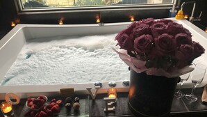 Una bañera llena de burbujas, un ramo de rosas moradas y una bandeja de bombones de chocolate.