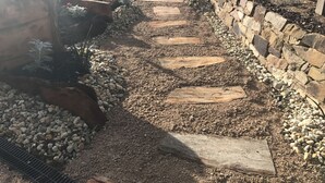 Un sendero de jardín con piedras y un muro de piedra, con un coche blanco al fondo.