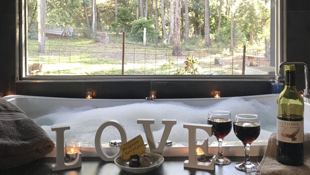 Una bañera con una toalla, una botella de vino, dos copas de vino tinto y velas en un alféizar.
