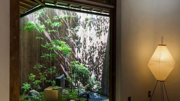Um quarto com piso de madeira, uma janela grande com vista para um jardim e um abajur.