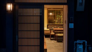 Uma entrada tradicional em estilo japonês, com uma porta deslizante, uma pequena área de estar ao ar livre com mesa e cadeiras, e um lampião à esquerda.