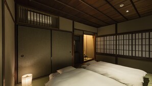 Quarto tradicional japonês com tatames, teto de madeira baixo e portas deslizantes shoji. Há duas camas com roupa de cama branca e uma pequena mesa com um abajur.