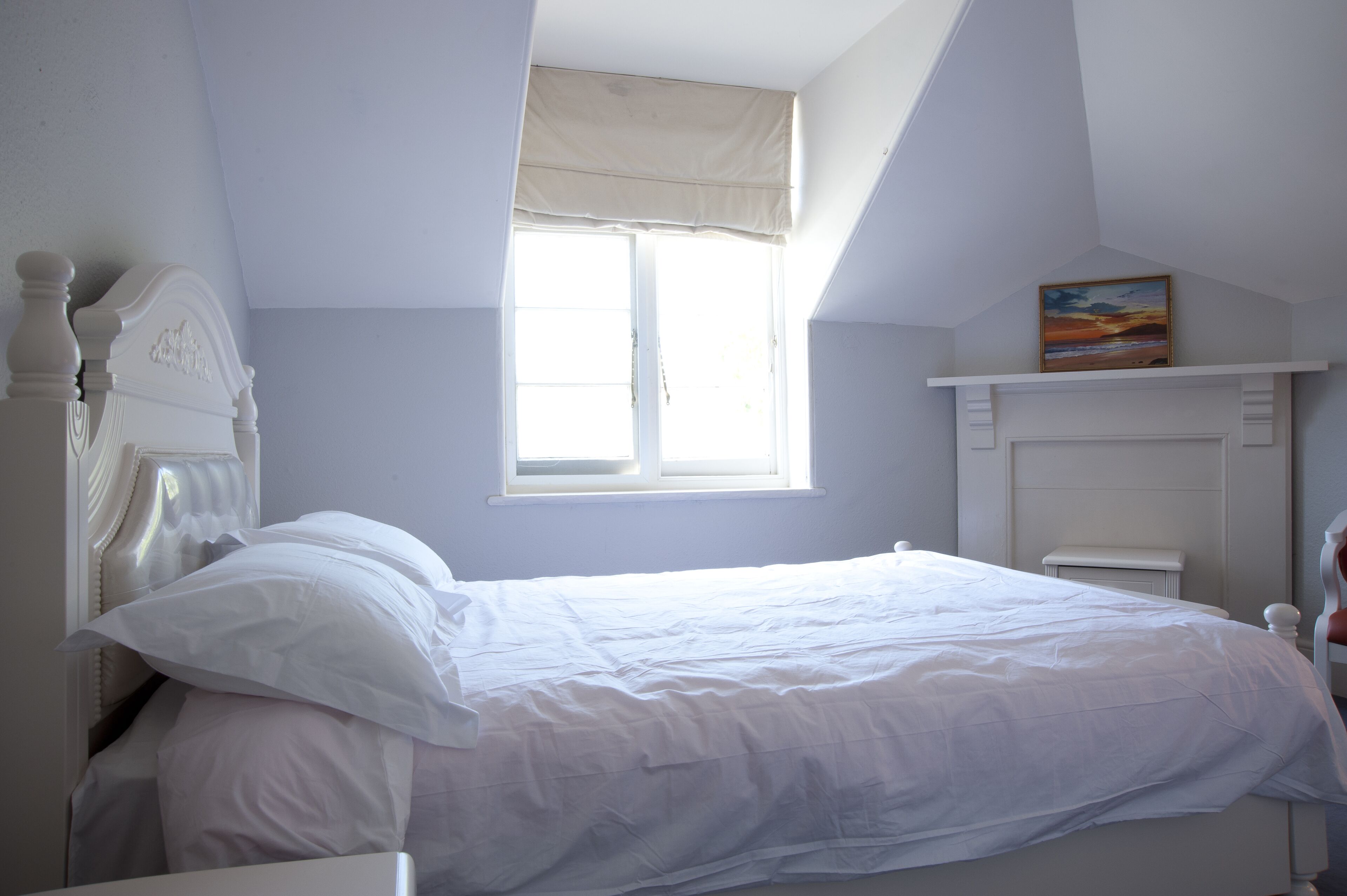 A bedroom with a large window, a bed with white bedding, a headboard, and a painting on the wall.