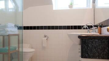 A bathroom with a glass shower enclosure, a white toilet, a black and white tiled floor, a sink with a black countertop, and a skylight above the toilet.
