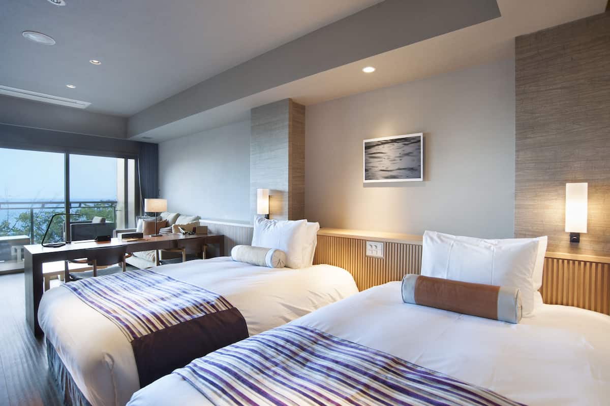A hotel room with two beds, a desk, a chair, a lamp, and a painting on the wall.