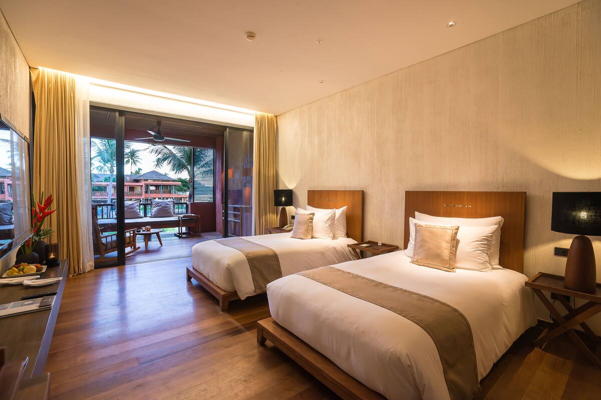 A hotel room with two beds, a desk, and a view of the outdoor area.