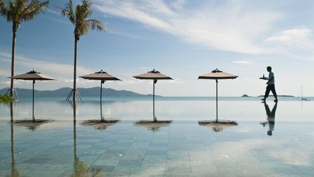 An infinity pool with four umbrellas, palm trees, and a person walking. - Hansar Samui Resort & Spa (Koh Samui)