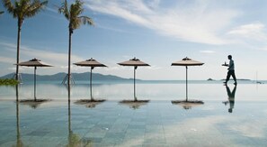 Ein Pool ohne sichtbare Kante mit vier Sonnenschirmen, Palmen und einer Person, die gerade geht.
