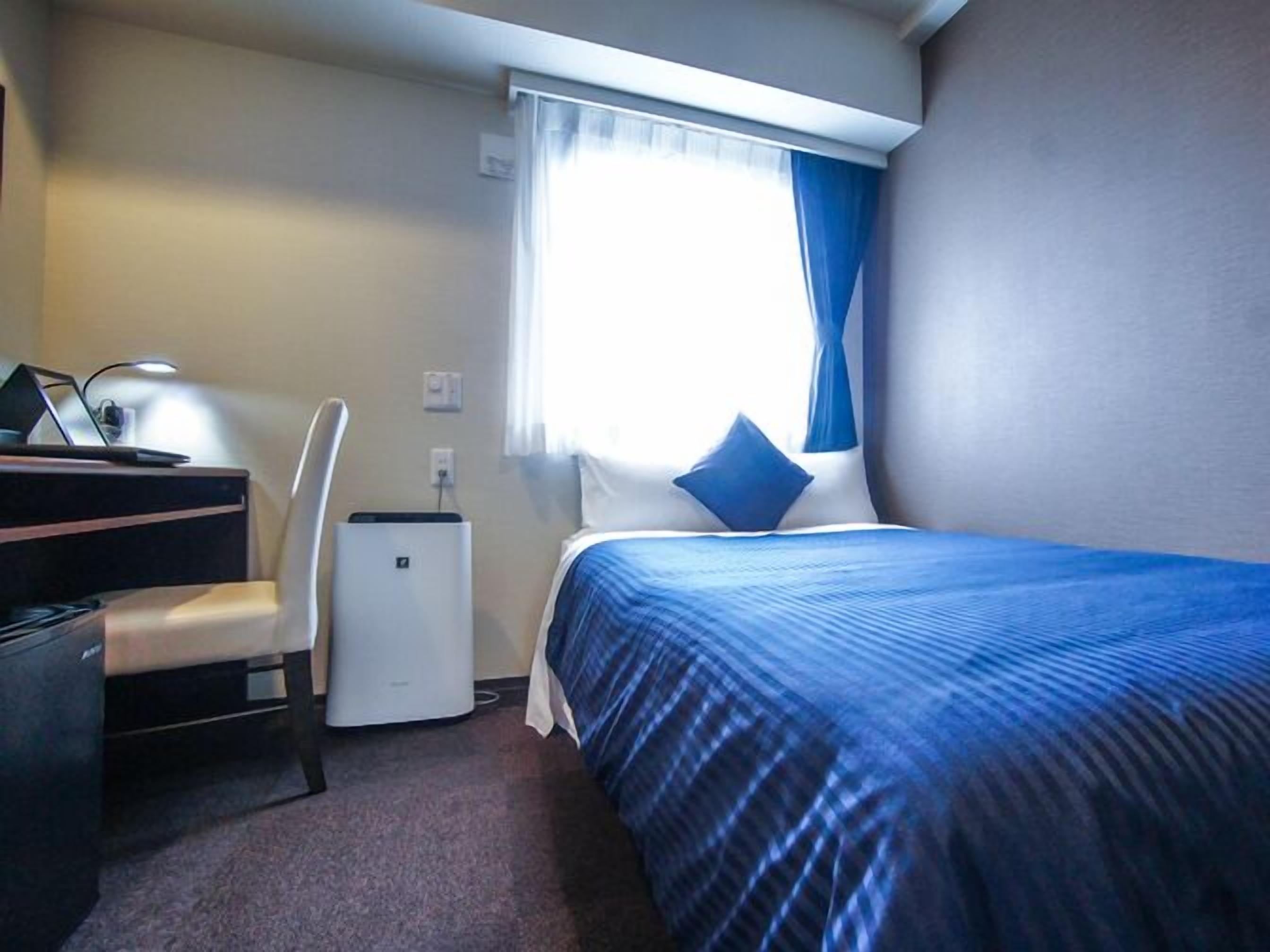 A hotel room with a bed, a desk, a chair, a small air conditioner, and a window with curtains.