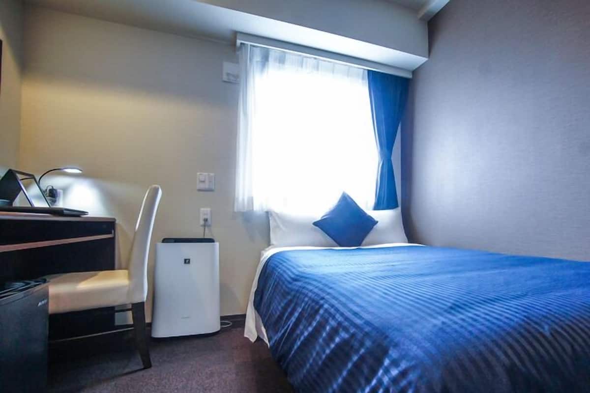 A hotel room with a bed, a desk, a chair, a small air conditioner, and a window with curtains.