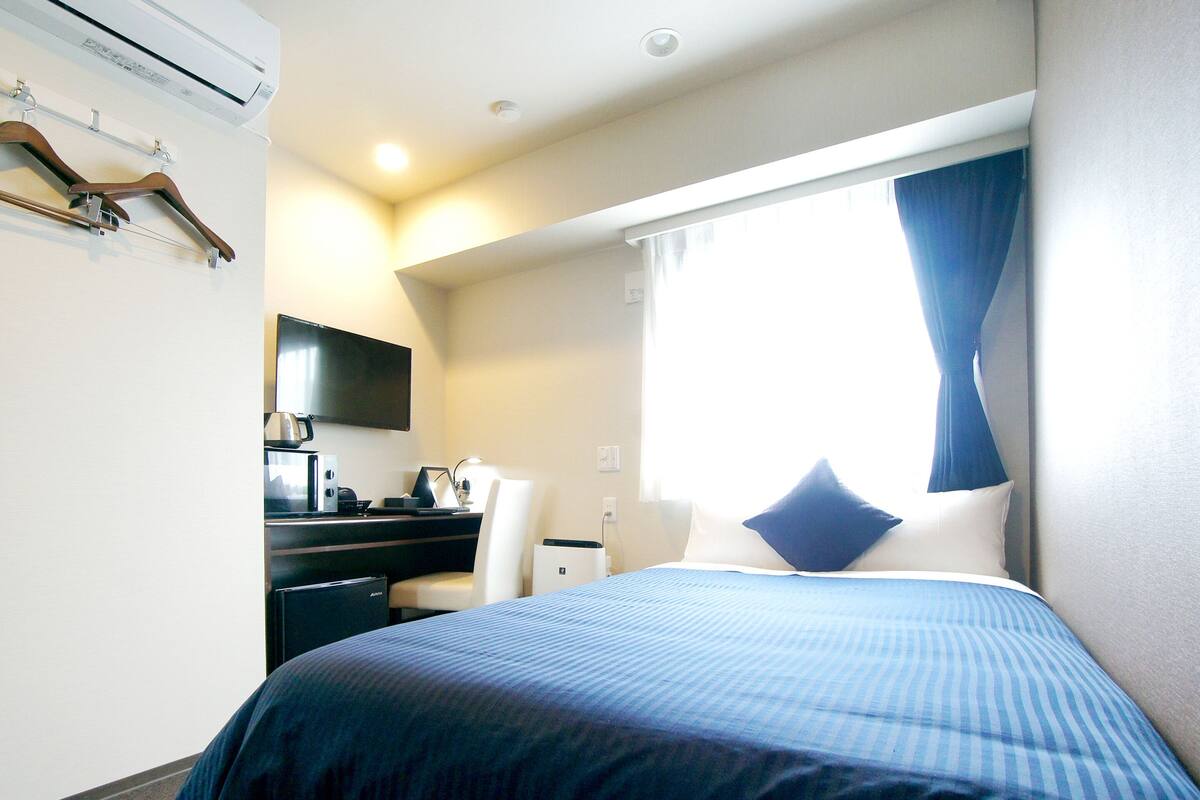 A hotel room with a bed, a desk with a chair, a flat-screen TV, and a window with curtains.