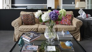 A spacious living room with high ceilings and exposed beams, a beige sofa, a coffee table with books and a vase of flowers, and a view of a kitchen and dining area through a large window.