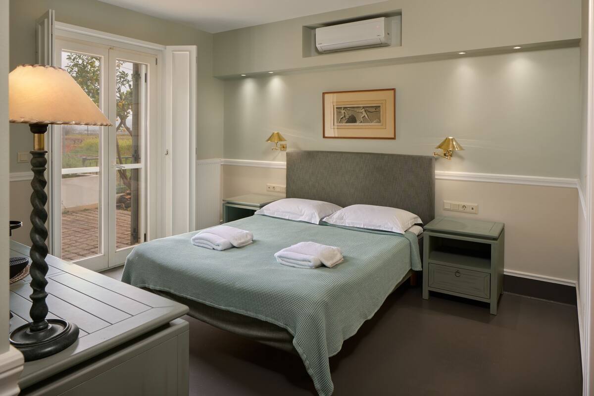 A neatly made bed with a green bedspread and two pillows, a bedside table with a lamp, and a small drawer unit.