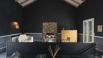 Une pièce aux couleurs sombres, avec une cheminée en pierre, un canapé blanc, un meuble en bois et une table en verre ornée de bougies et de bâtonnets d’encens.