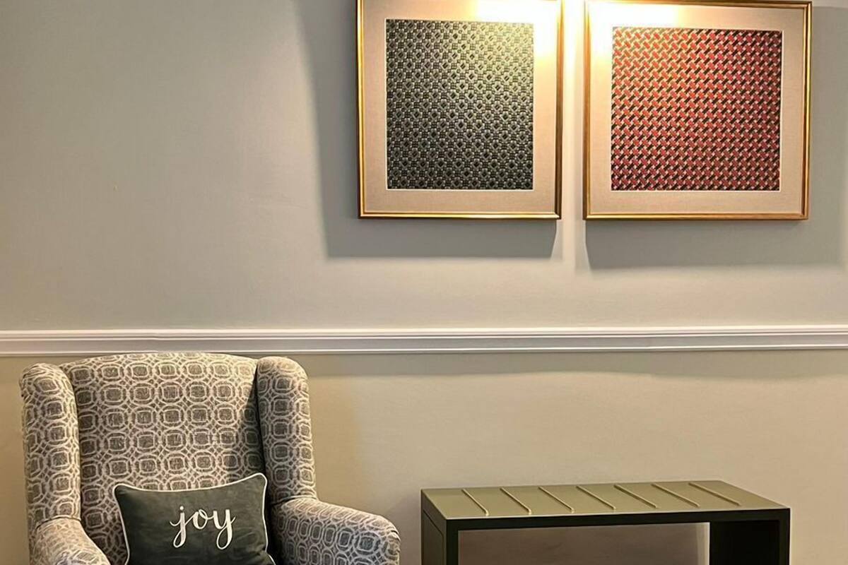A living room with a patterned armchair, a matching ottoman, a small side table, and two framed artworks on the wall.