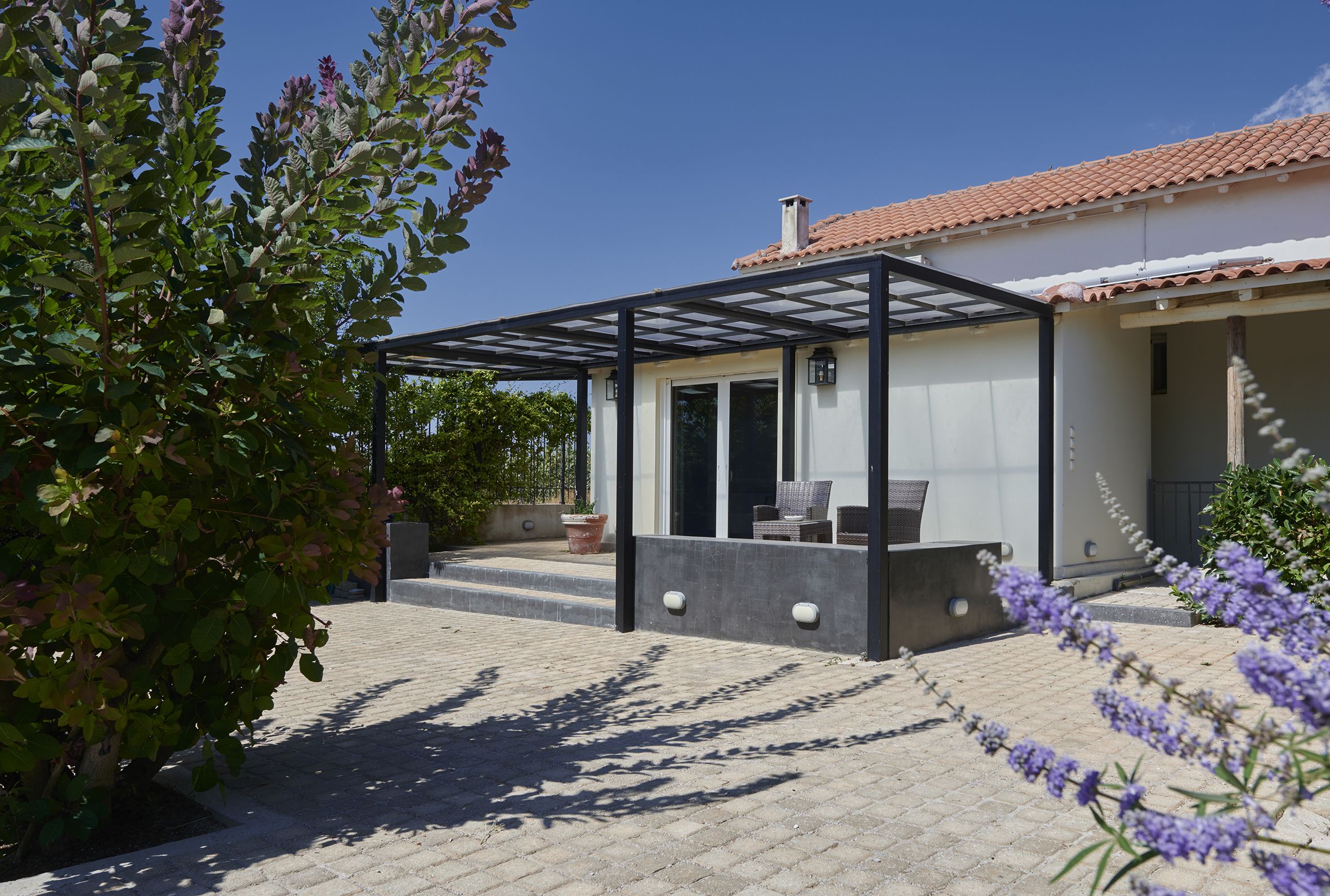 Un espacio de estar al aire libre cubierto, con una pérgola, una planta en maceta y vistas a una casa con tejado de tejas.