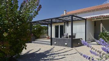 Un espacio de estar al aire libre cubierto, con una pérgola, una planta en maceta y vistas a una casa con tejado de tejas.