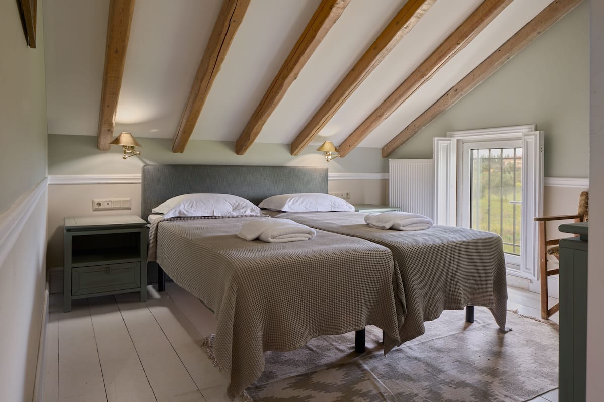 A bedroom with two beds, wooden beams on the ceiling, bedside lamps, and a window with a view.