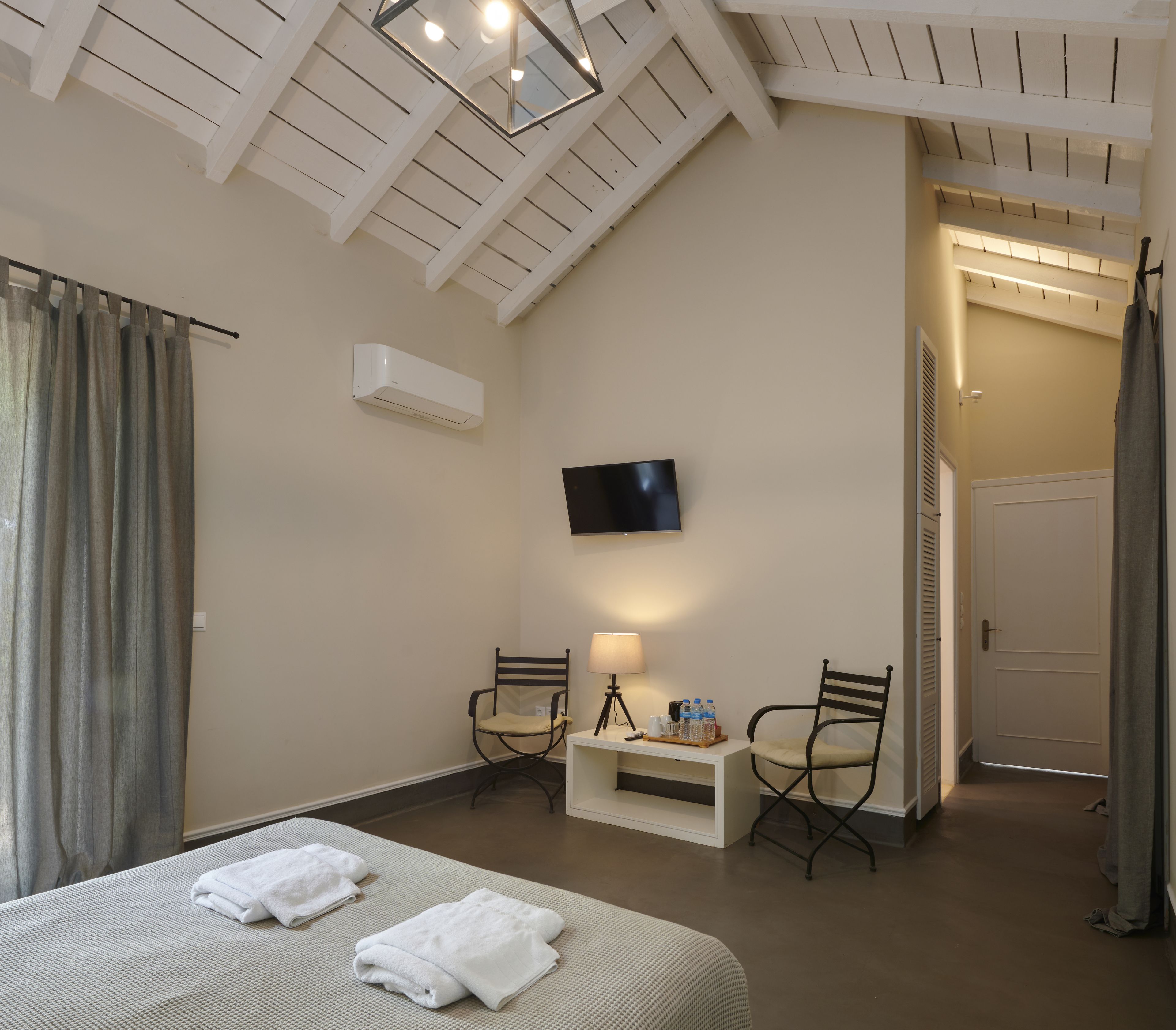A hotel room with a bed, two chairs, a TV, a desk with a lamp, and an air conditioning unit.