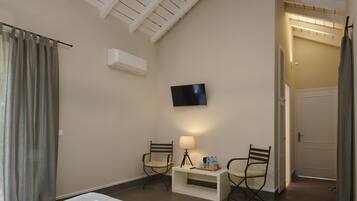 A hotel room with a bed, two chairs, a TV, a desk with a lamp, and an air conditioning unit.