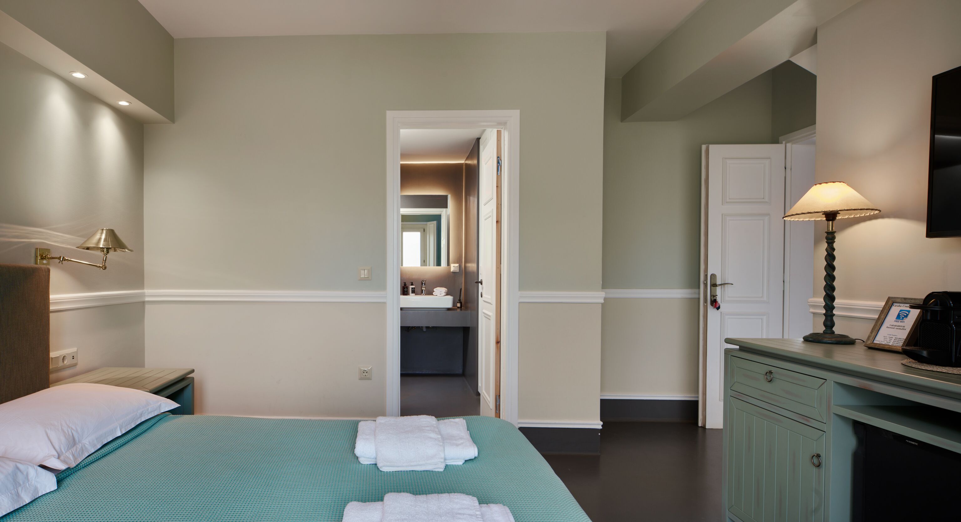 Een hotelkamer met een bed, een bureau met lamp, een kledingkast en een badkamer zichtbaar door een geopende deur.