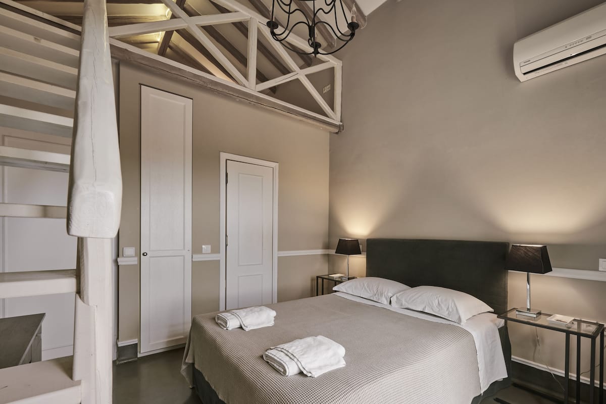 A bedroom with a bed, two bedside tables, a nightstand, a wall-mounted air conditioner, and a door.