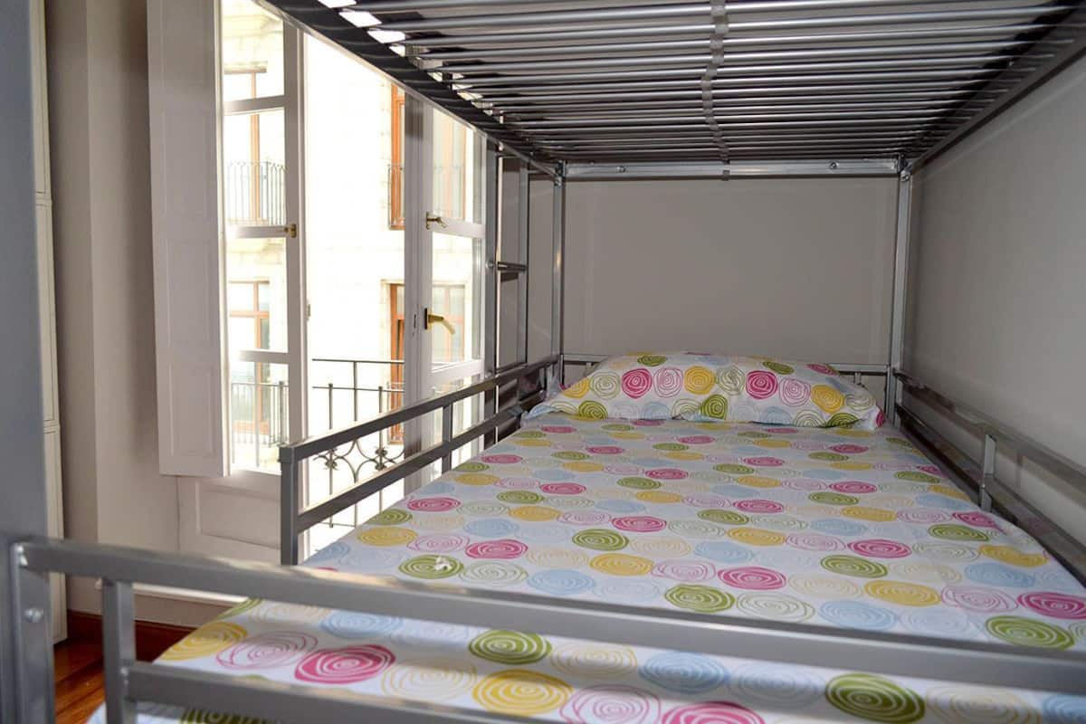 A small, single-bed room with a metal bed frame, a colorful bedspread, and a window with a metal railing.