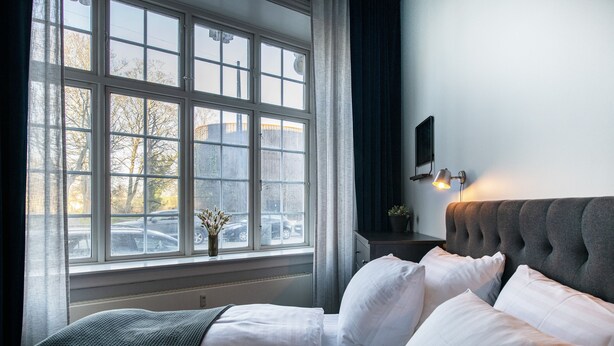 Quarto com uma janela grande, cama com roupa de cama branca, cabeceira estofada cinza e mesa de cabeceira com abajur.