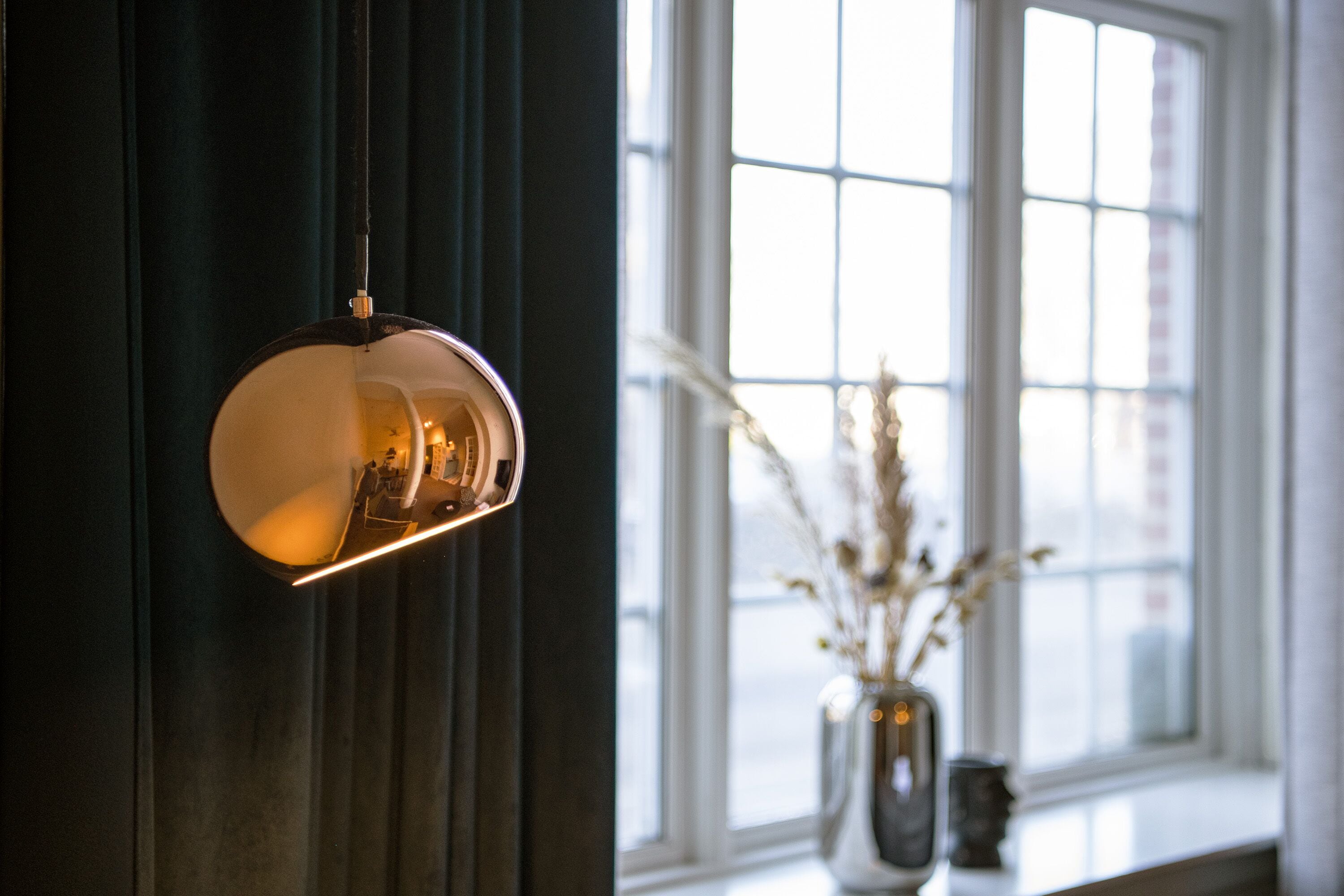 A pendant light fixture in a room with a window and a vase with dried plants.
