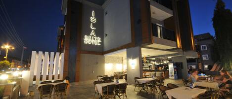 An outdoor dining area with tables and chairs, a bar, and a neon sign for the Isla Suite Hotel.