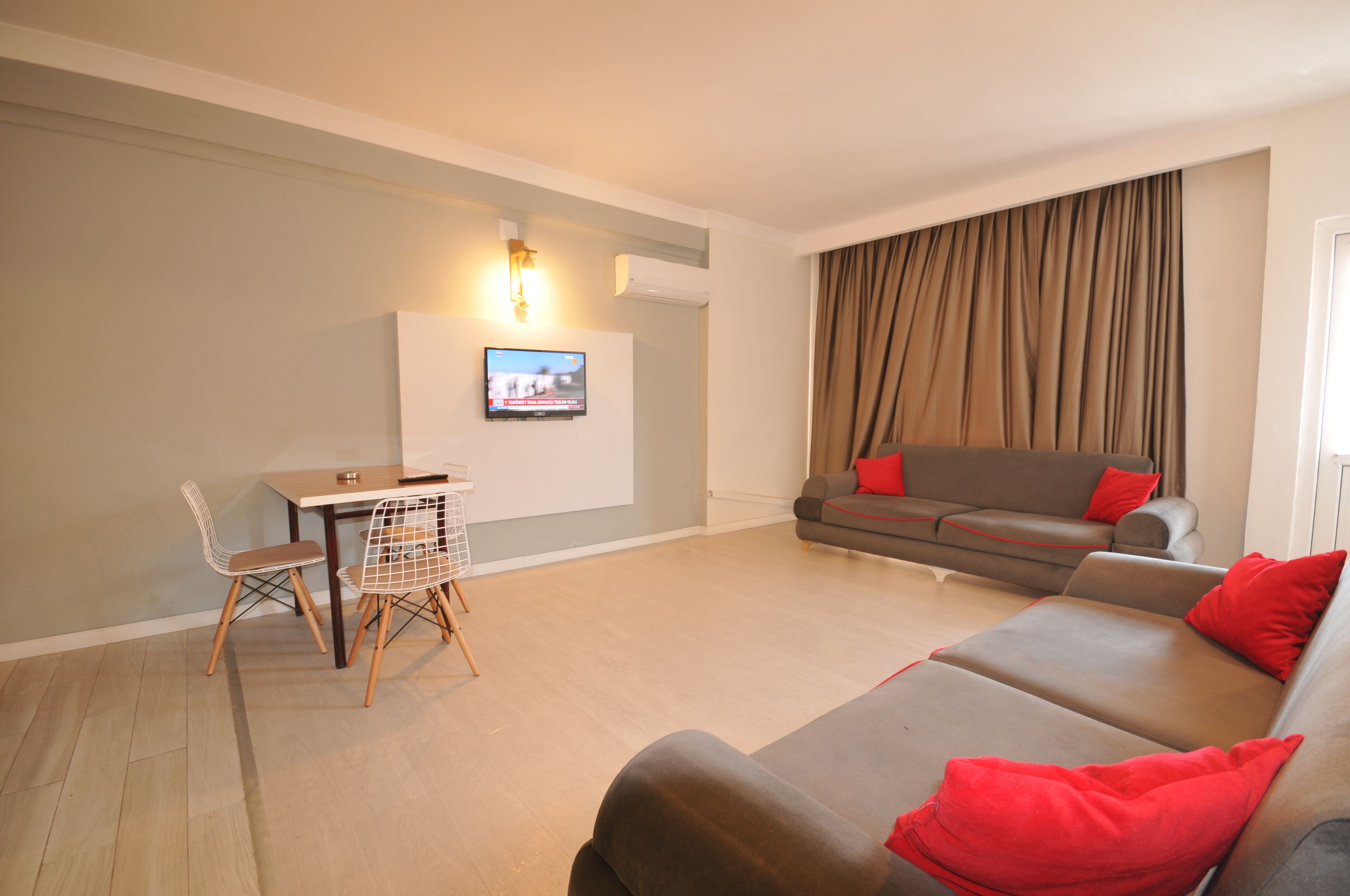 A modern living room with a sofa, a dining table with chairs, a flat-screen TV mounted on the wall, and a large window with curtains.