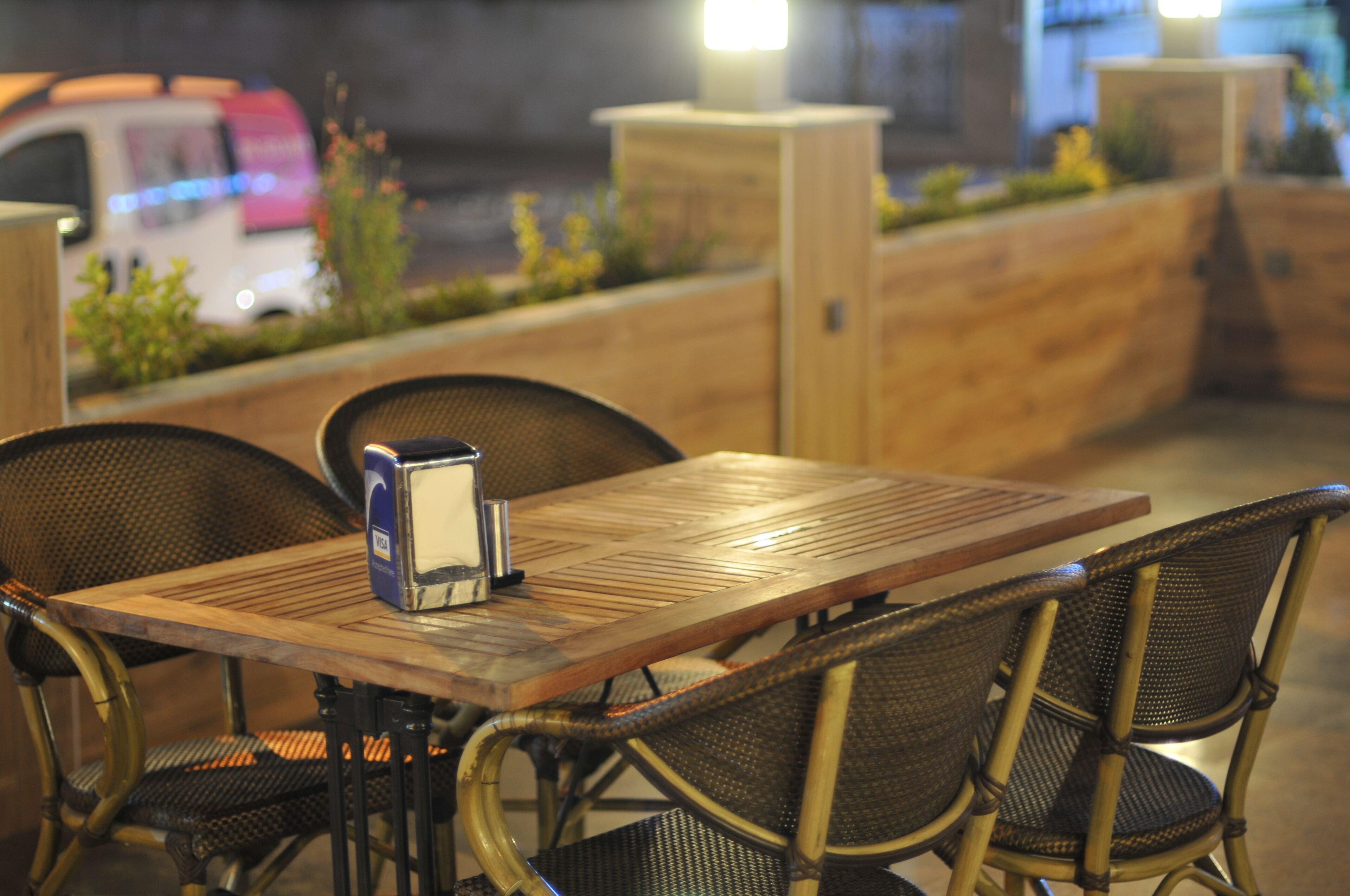 An outdoor seating area with a wooden table and wicker chairs, a coffee machine, and a wooden partition with plants.