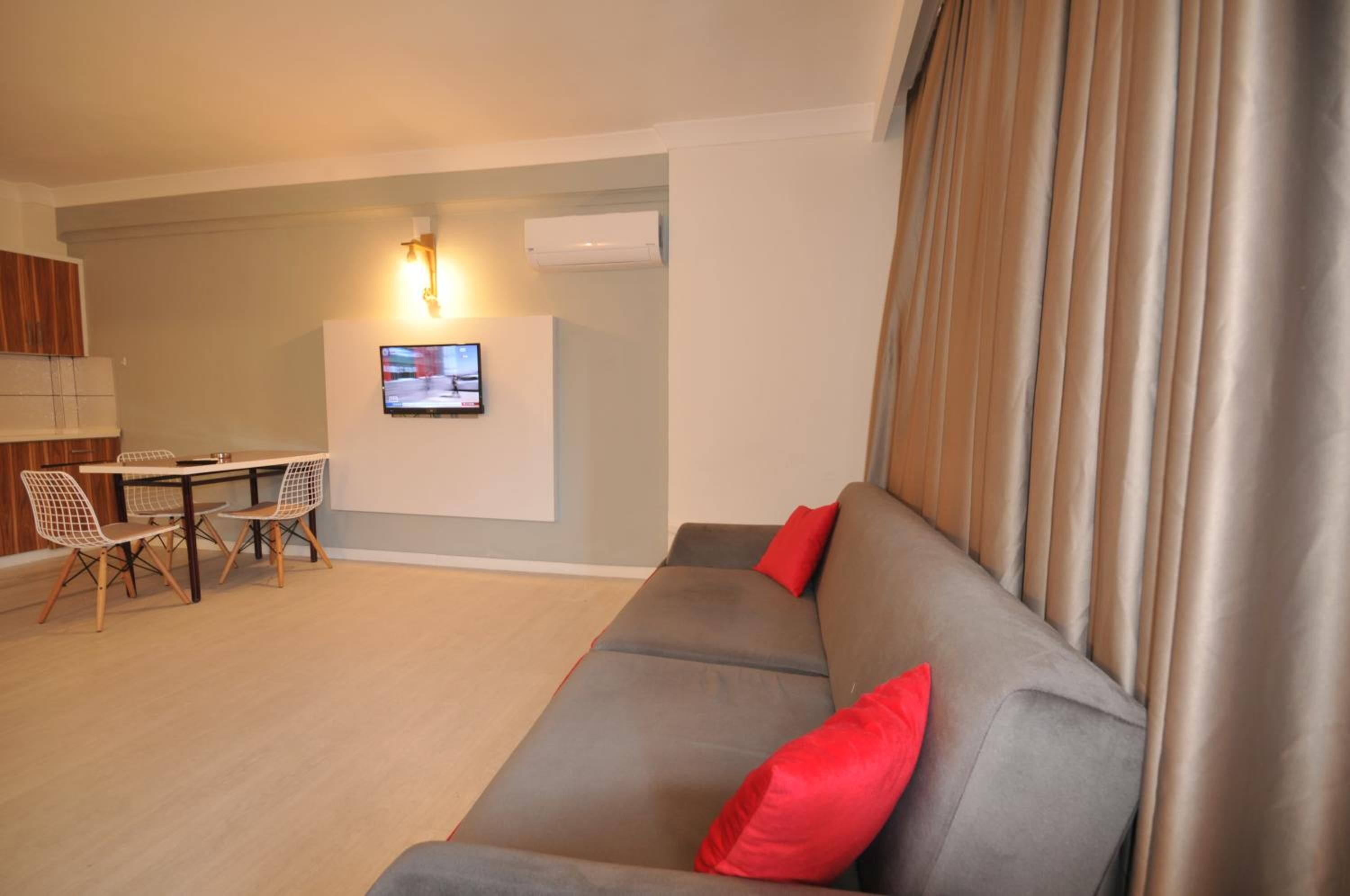 A modern living room with a grey sofa, red cushions, a dining table with chairs, and a flat-screen TV mounted on the wall.