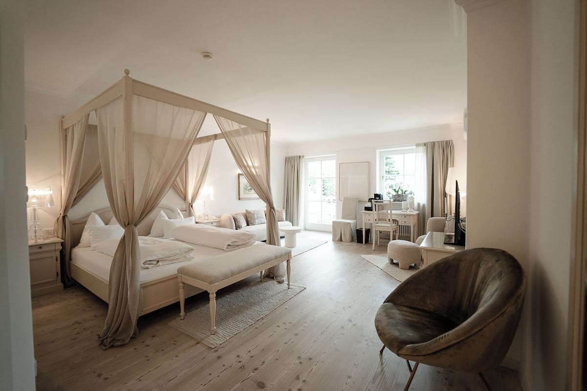 A bedroom with a four-poster bed, a sitting area with a chair, a desk, and a small table.