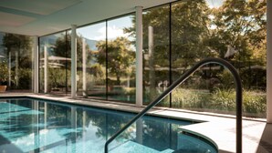 An indoor swimming pool with clear glass walls, a metal handrail, and a view of trees and mountains outside. - Hotel Hanswirt (Parcines)