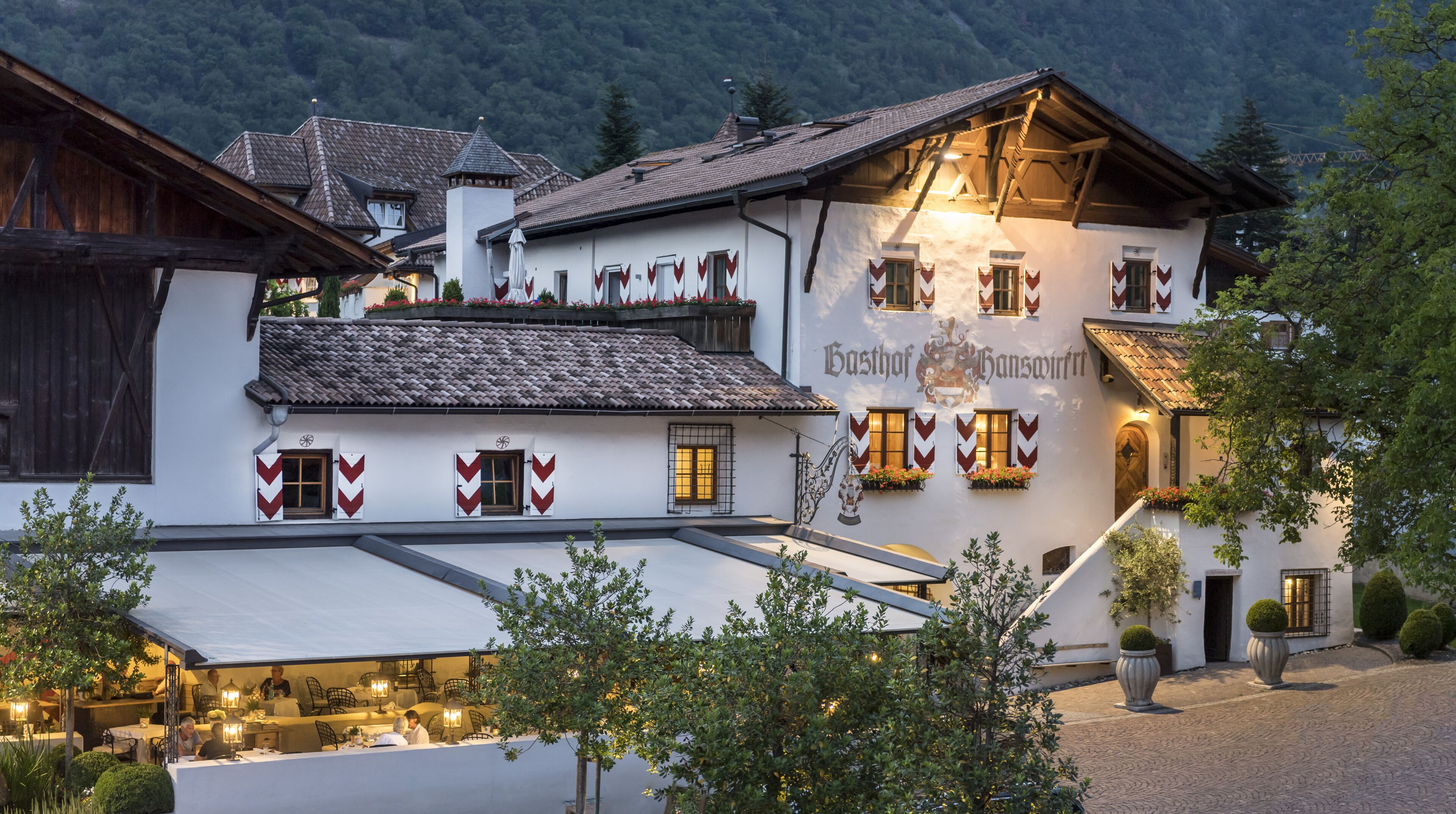 Ein traditionelles europäisches Gebäude mit einem Schild, auf dem "Gasthof Hanswirt" steht. Es hat eine weiße Fassade, hölzerne Balkone und ein Ziegeldach.