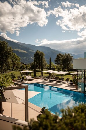 A swimming pool with lounge chairs and umbrellas, surrounded by greenery and mountains in the background. - Hotel Hanswirt (Parcines)